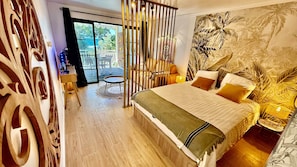 Ein Schlafzimmer mit einem Bett, einem kleinen runden Tisch, einem Sessel, einer Wand mit Dekoration und Blick durch eine Glastür.