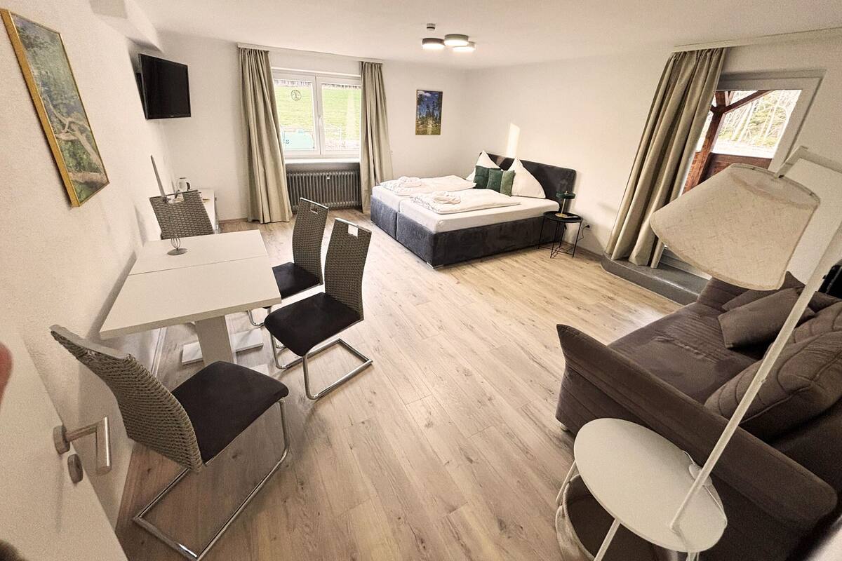 A hotel room with a bed, a desk with a chair, a sofa, a TV mounted on the wall, and a window with curtains.