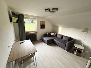 A room with a sectional sofa, a small table, a radiator, a TV, a lamp, and a window with curtains. - pfefferHÄUSL Pension - Camping (Regen)