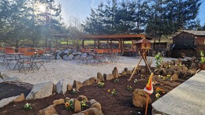 Un espacio al aire libre con mesas y sillas, una estructura de madera y un jardín con rocas y flores.