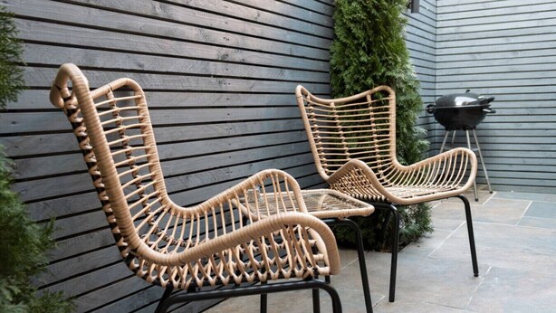 Zwei Korbstühle mit Metallgestellen, ein kleiner Tisch und ein Grill befinden sich auf einer Terrasse mit grauer Holzverkleidung und Steinboden.
