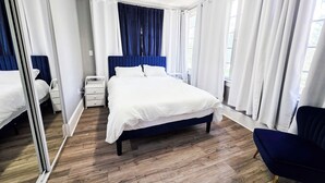 A bedroom with a bed, a nightstand, a chair, a mirror, and a window with curtains.