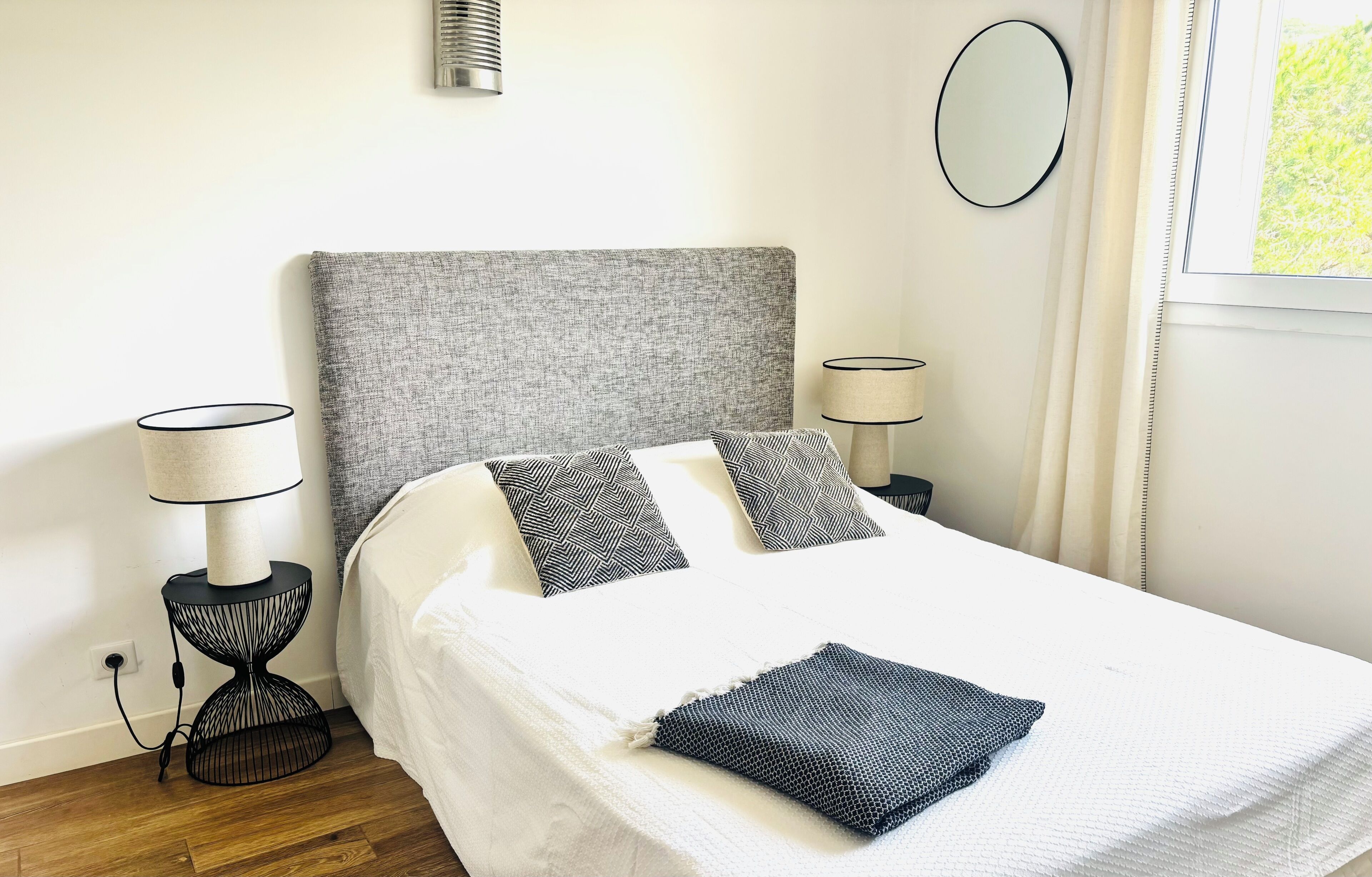 A neatly made bed with a grey headboard, two bedside lamps, a round mirror, and a window with curtains.