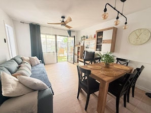 A modern living room with a sofa, dining table, chairs, a ceiling fan, and a large clock on the wall.
