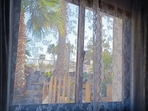 A window with patterned curtains, a view of a fence, and palm trees.