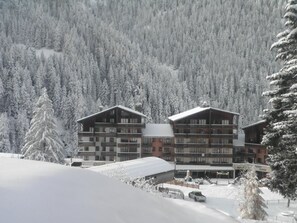 A multi-story hotel with balconies, surrounded by snow-covered trees and a mountainous backdrop. - 20m from resort center, 5th floor, balcony, tv, ski locker, 21m², Valfréjus (Valfréjus)