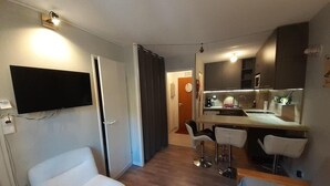 Um espaço de estar compacto com televisão na parede, cozinha compacta com banquetas altas e uma porta que dá para outro quarto.