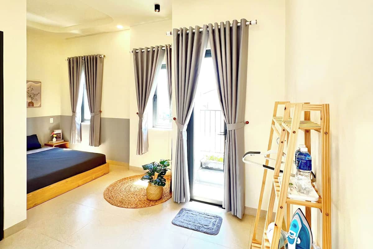 A modern room with a bed, a small table with a plant, a wooden shelf with towels, and a view of the outside.