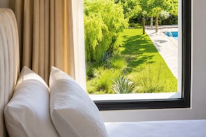 Una habitación con cama, almohadas blancas y ventana con vistas a un jardín cuidado y una piscina.