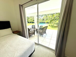 A hotel room with a bed, a small table, wicker chairs, and a view of a swimming pool and garden.