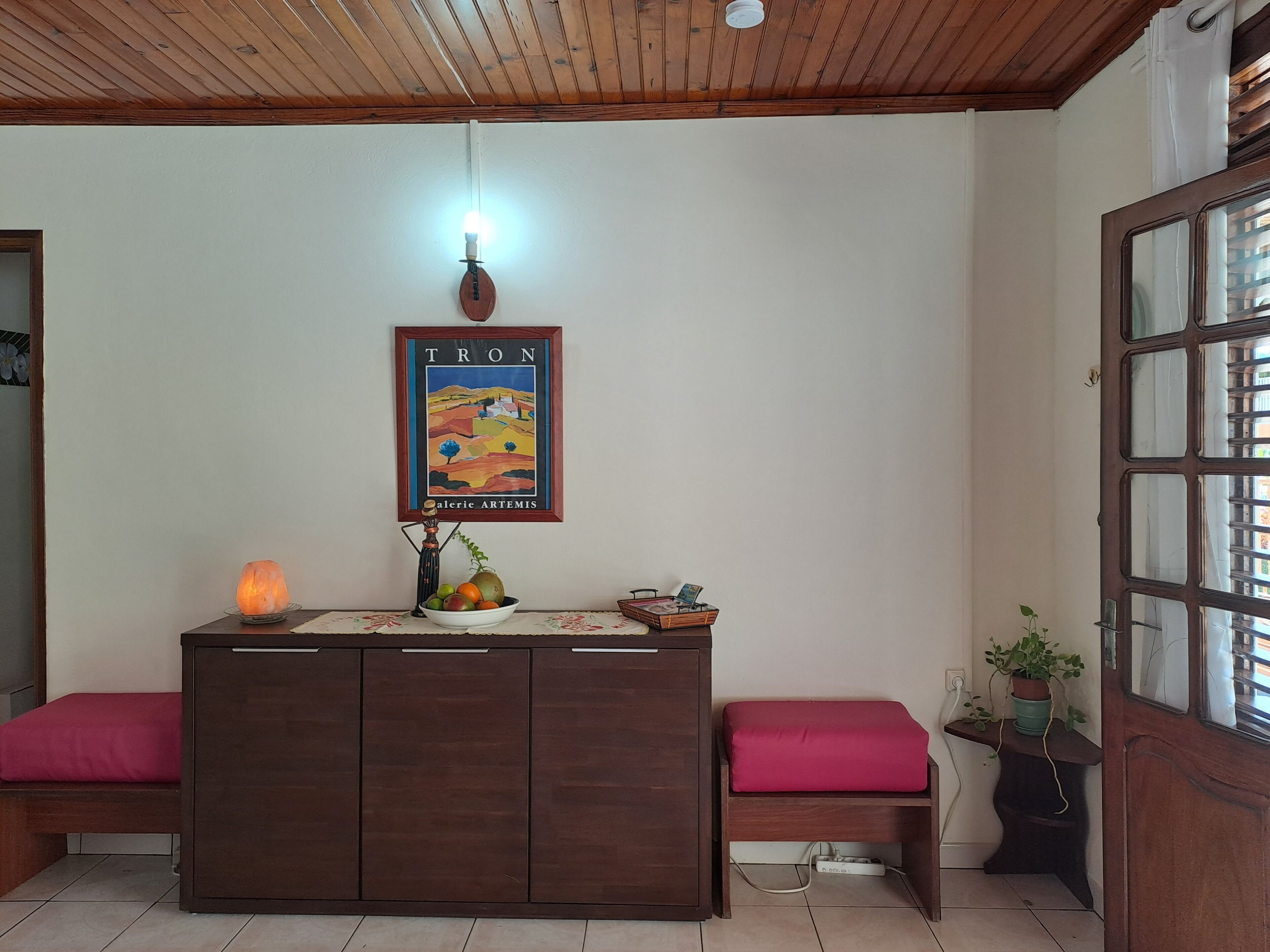 A room with a wooden ceiling, a framed poster on the wall, a small table with a fruit bowl, a heater, and a bench with a cushion.