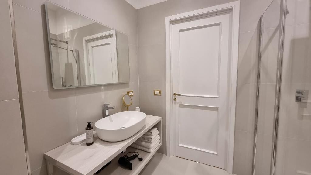 A modern bathroom with a white sink, a large mirror, a door, and a shower area.
