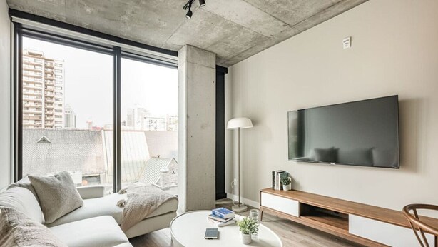 A modern living room with a sofa, coffee table, TV, and a view of the city through large windows. - Modern Urban Retreat with Skyline Panorama (Montreal)
