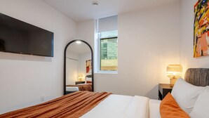 A hotel room with a bed, a TV mounted on the wall, a window with a view, and a nightstand with a lamp. - Vintage Moderne – Old MTL Retreat (Montréal)