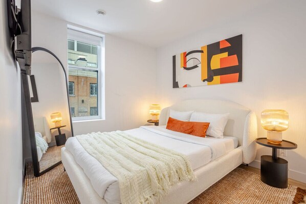 A bedroom with a bed, two bedside tables, a mirror, a window, and a wall art piece. - Historic Hideaway – Bright Apartment in Old Montreal (Montréal)