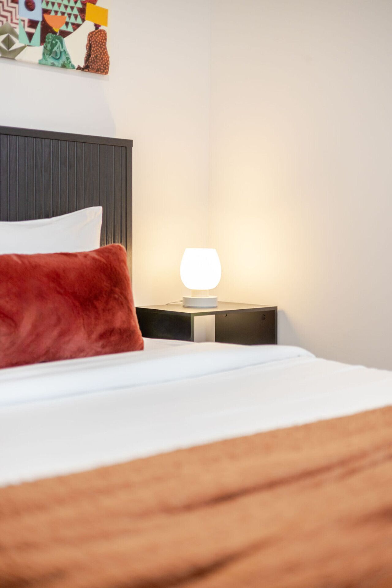 A neatly made bed with a white and brown duvet, a red pillow, and a bedside table with a white lamp.