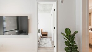 Ein modernes Zimmer mit einem an der Wand montierten Flachbildfernseher, einem hölzernen Bett, einer großen Topfpflanze und einem Flur, der in ein weiteres Zimmer führt.