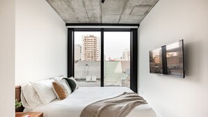 Une chambre à coucher comprenant un lit, une table de chevet, un téléviseur fixé au mur et une grande fenêtre donnant sur la ville.