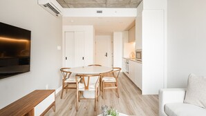 Un salon moderne avec une table de salle à manger ronde, des chaises en bois, un téléviseur à écran plat fixé au mur, un coin cuisine avec des meubles blancs et un canapé de couleur claire.