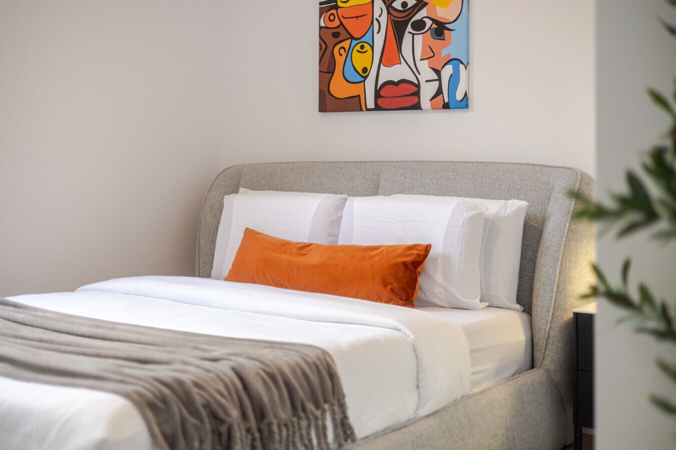 A neatly made bed with white and orange pillows, a grey headboard, and a colorful abstract painting on the wall.