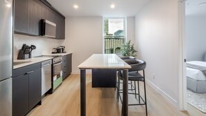 A modern kitchen with dark cabinetry, a stainless steel refrigerator, and a dining area with a table and chairs. - Stylish Downtown Retreat (Montreal)
