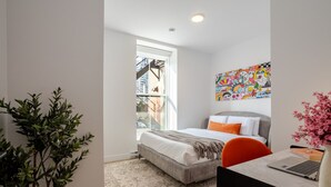 Een moderne slaapkamer met een bed, een bureau met een laptop, een potplant en een kleurrijk kunstwerk aan de muur.