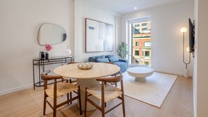 Ett modernt vardagsrum med ett runt matbord, två trästolar, en blå soffa, en stor abstrakt målning och en utsikt över byggnader genom ett fönster.