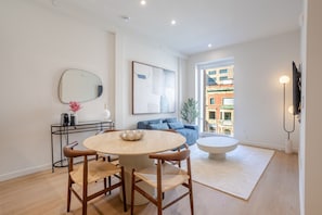 A modern living room with a round dining table, two wooden chairs, a blue sofa, a large abstract painting, and a view of buildings through a window. - Modern Retreat with Chic Design in Downtown MTL (Montreal)