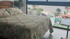 Un dormitorio con una cama grande, vistas a la ciudad y un balcón con una silla de macramé.