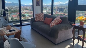 Una sala de estar con sofá, una mesa de centro y vistas a un paisaje urbano a través de amplios ventanales.