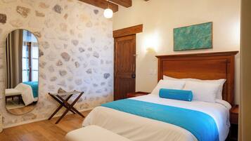 A bedroom with a stone wall, wooden ceiling beams, a bed with white and blue bedding, a wooden nightstand, a mirror, and a painting on the wall.