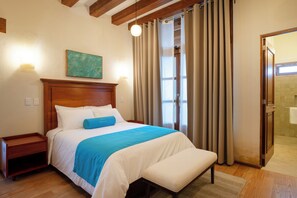 Habitación de hotel con cabecera de madera, cama con ropa de cama blanca y azul, mesita de noche, lámpara de pared y zona de ducha con cortinas.