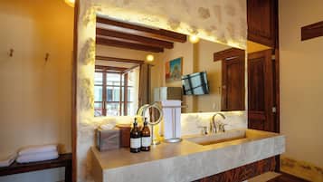 Un baño con pared de piedra, vigas de madera, un espejo grande, un lavamanos y un televisor montado en la pared.