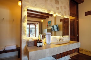 Une salle de bain avec un mur en pierre, des poutres en bois, un grand miroir, un lavabo et un téléviseur fixé au mur.