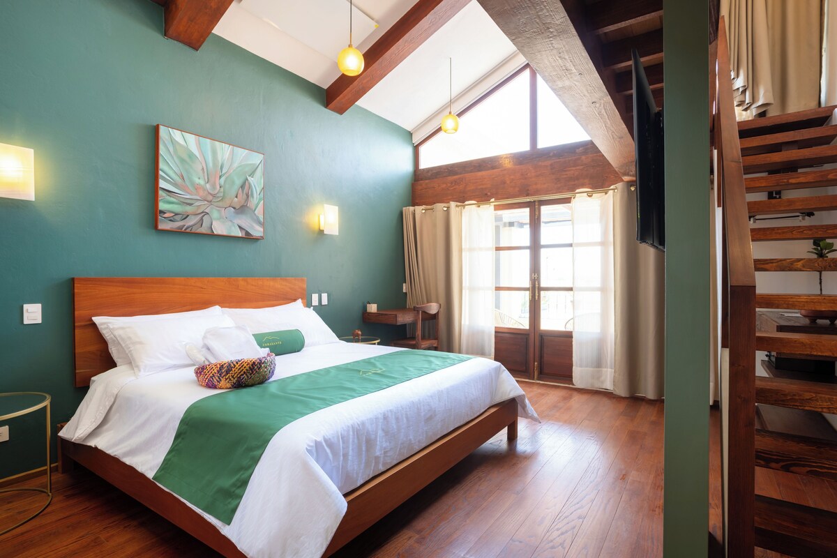 Quarto de hotel com cama de madeira, manta verde, quadro na parede e janela com cortinas.