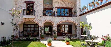Un edificio de dos pisos con fachada de piedra, varios balcones con plantas en macetas y espacios con mesas al aire libre.