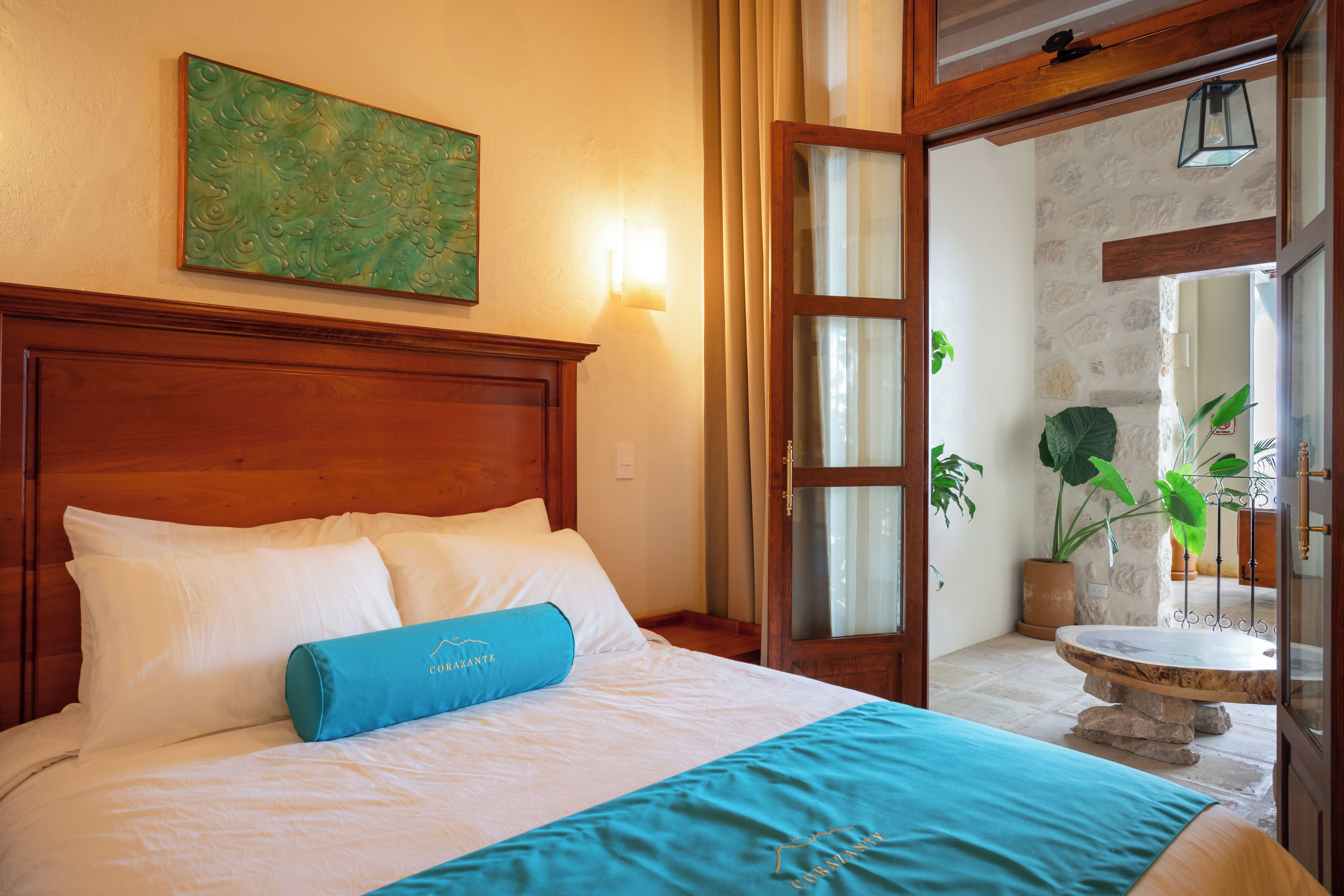 Habitación de hotel con cabecera de madera, cama con ropa de cama blanca, almohada decorativa azul, cuadro abstracto verde en la pared, y vista a una pared de piedra y plantas a través de una puerta abierta.