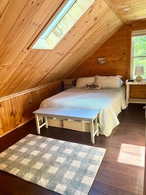 Ein Schlafzimmer mit schrägen Wänden, einem Bett mit gemusterten Betttextilien, einem Nachttisch mit Lampe und einer kleinen Bank.