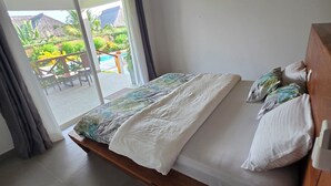 Ein Schlafzimmer mit Bett, Holz-Kopfteil, Fenster mit Vorhängen und Blick auf Pool und Grünfläche.