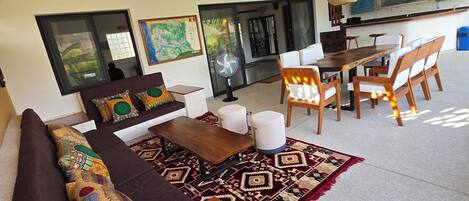 A modern living room with a wooden ceiling, a sofa with patterned cushions, a dining area with chairs, and a large rug.