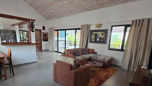 Ein geräumiger Wohnbereich mit einer Couch, einem Couchtisch und einem Esstisch mit Stühlen. Ein großes Fenster lässt natürliches Licht herein und bietet einen Blick ins Freie.