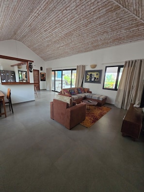 A spacious living area with a sofa, a coffee table, and a dining table with chairs. There is a large window providing natural light and a view of the outdoors.