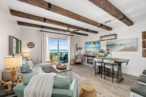 Un salon avec un canapé, une table à manger et des chaises, un ventilateur de plafond et une vue sur l’océan par une grande fenêtre.