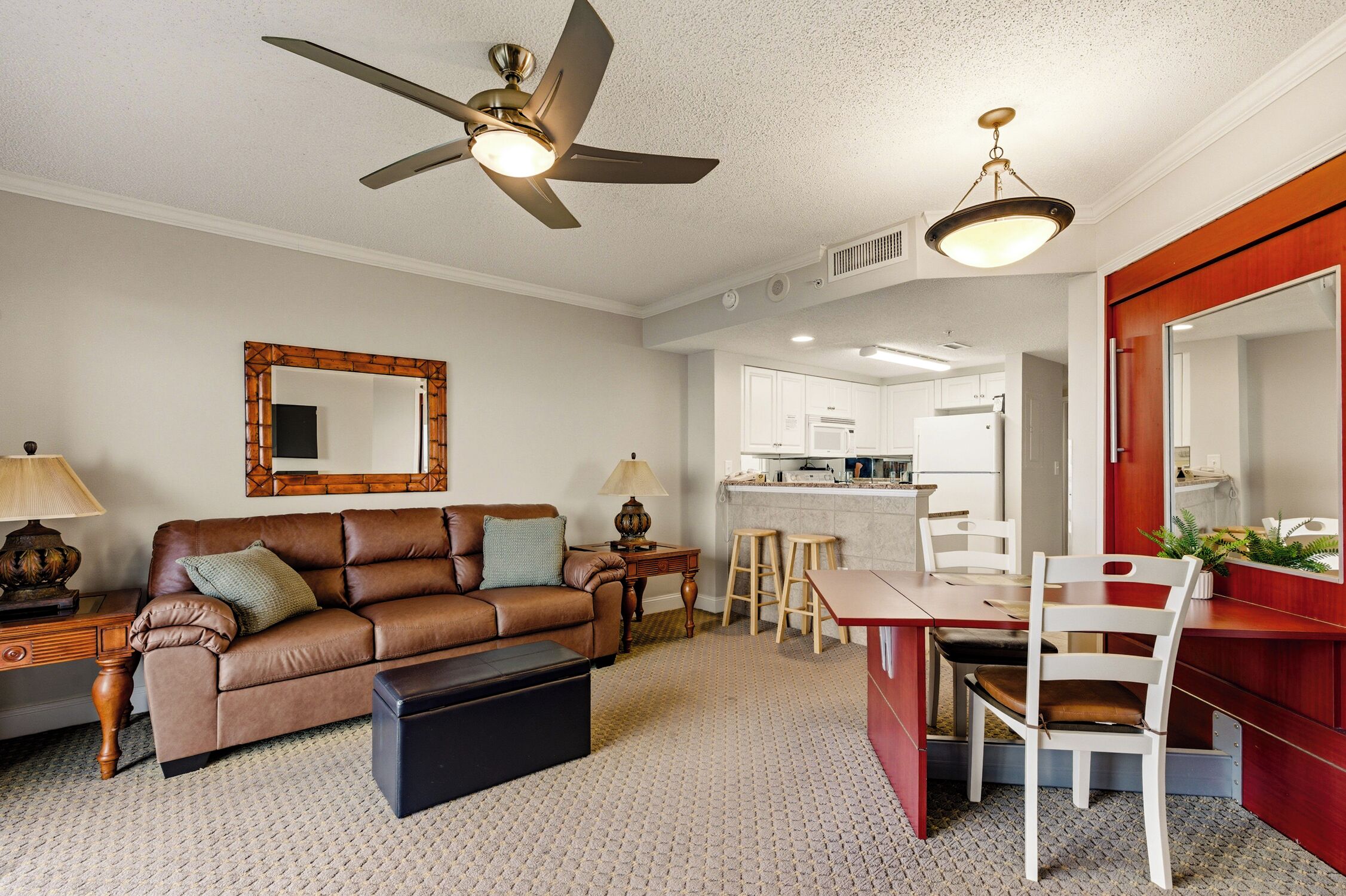 A living room with a sofa, a coffee table, a dining table with chairs, a kitchen in the background, and a ceiling fan.
