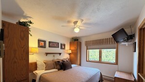 Quarto com cama, ventoinha no teto, TV fixada na parede e janela com vista para árvores.