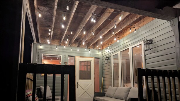 An outdoor porch with a wooden deck, a cushioned seating area, and string lights. - Steps from the Shore, Modern Beachy Getaway (Dunnottar)