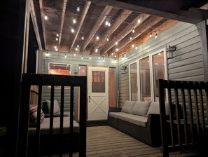 An outdoor porch with a wooden deck, a cushioned seating area, and string lights. - Steps from the Shore, Modern Beachy Getaway (Dunnottar)