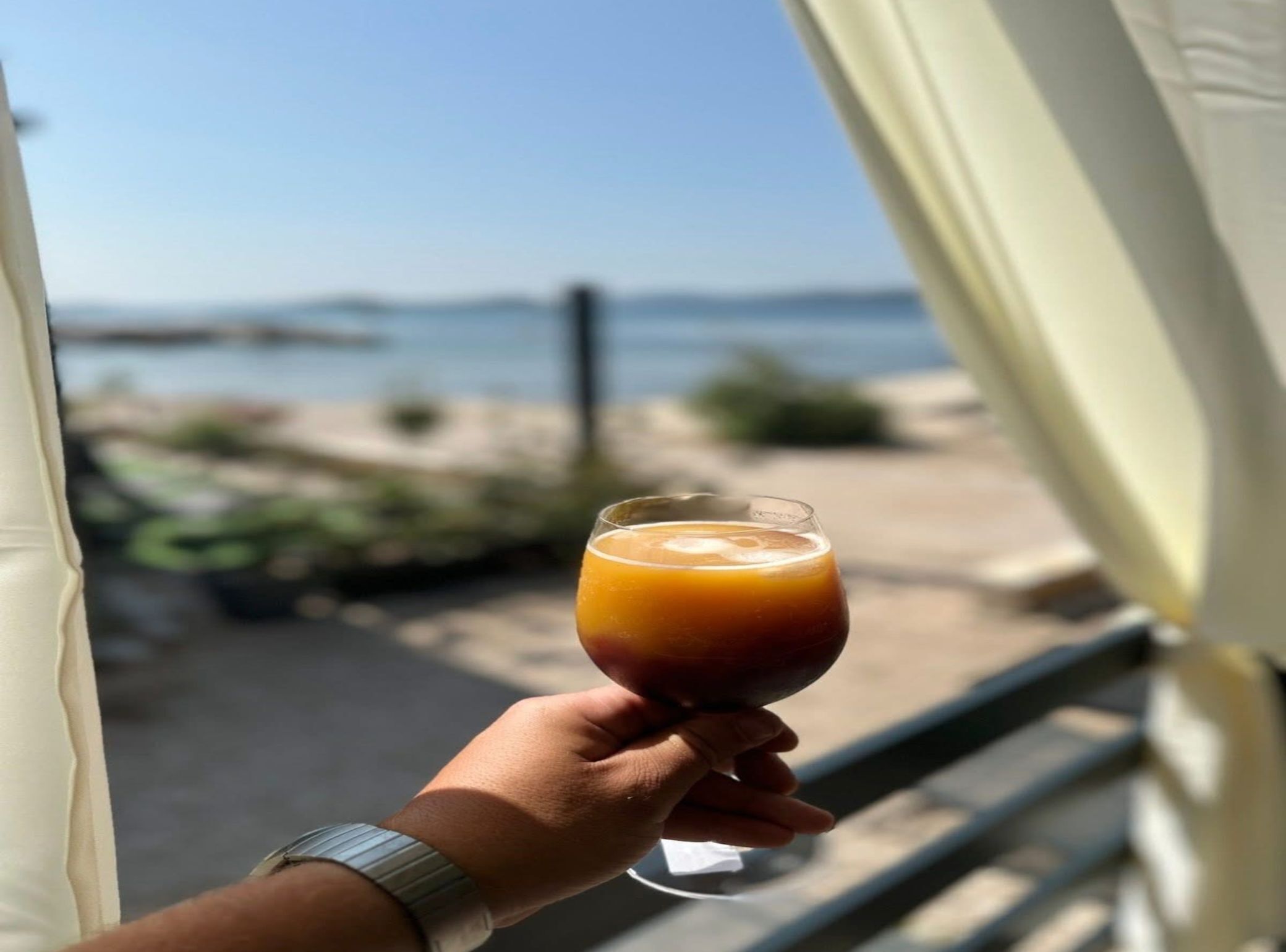 A hand holding a glass of orange juice, with a view of the beach and ocean through a window.