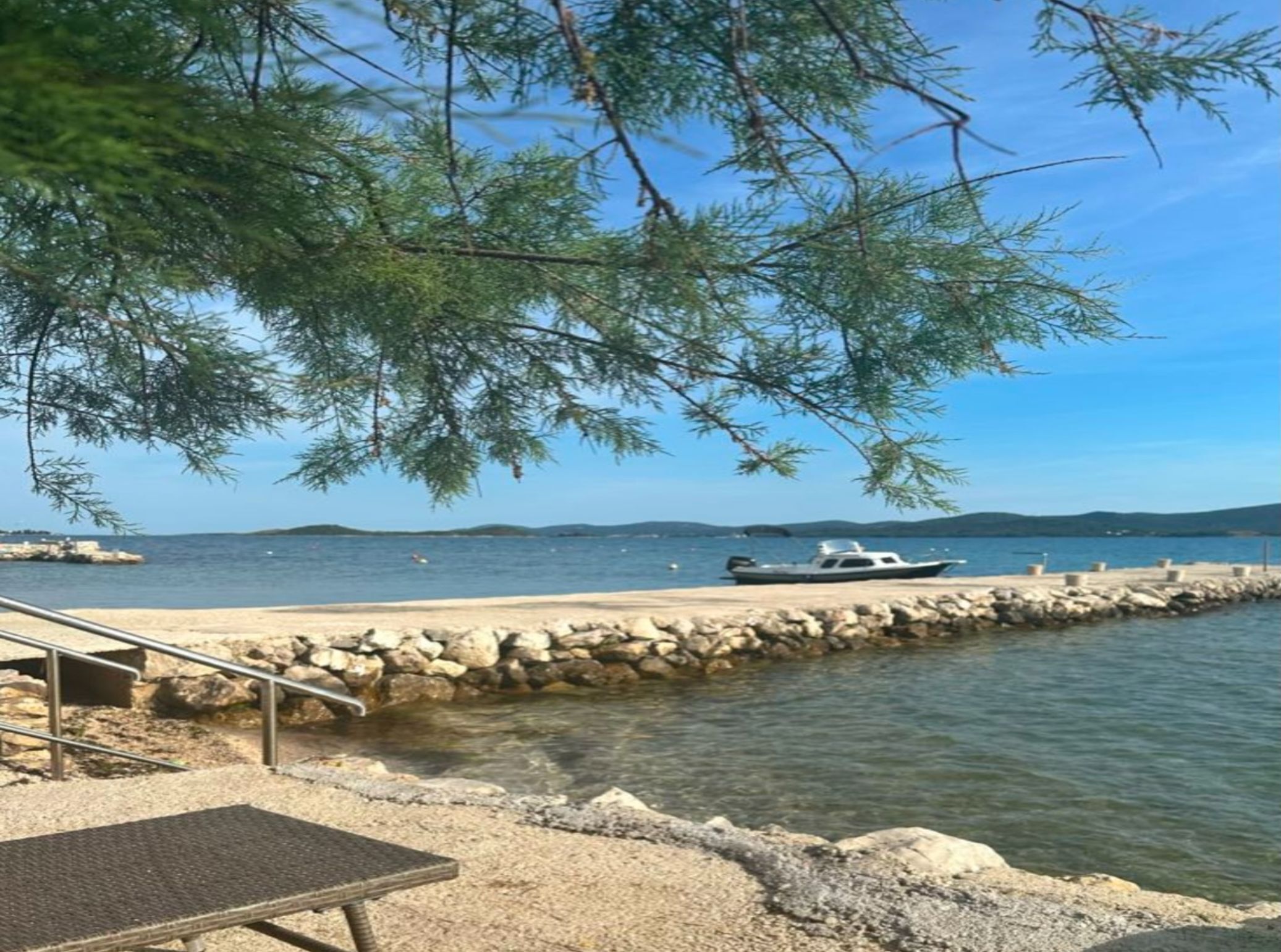 A waterfront area with a boat docked, a rocky shoreline, and a metal bench.