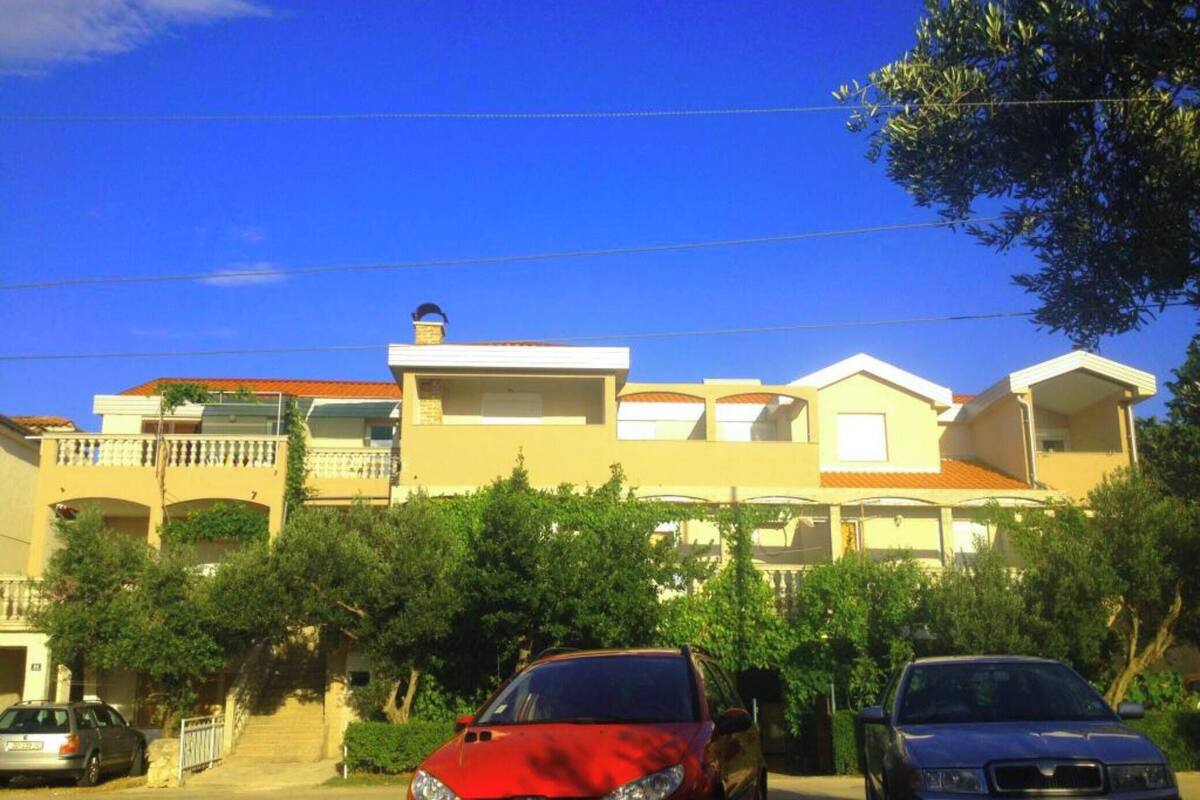 A residential building with multiple balconies, a red car parked in front, and a clear blue sky.